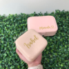 Una mano sostiene un Joyero personalizado de cuero sintético mini de color rosa con la etiqueta "Isabel" sobre otro estuche con la etiqueta "Marcela", ambos descansan sobre hojas verdes.
