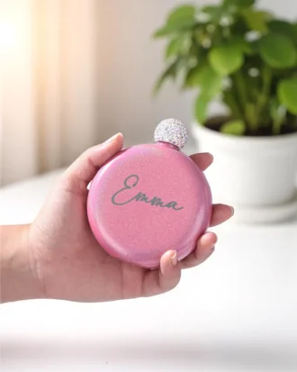 Una mano sostiene una Licorera personalizada de acero inoxidable 148ml 5oz rosa y redonda con la etiqueta Emma junto a una maceta verde.