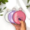 Una mano sujeta tres brillantes frascos con tapón joya Licorera personalizada de acero inoxidable 148ml 5oz en plata, morado y rosa -cada uno etiquetado Aurora- para dar un toque chic y personalizado.