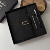 A Set libreta con broche magnético contorno metálico y bolígrafo en caja, color negro, junto a un portavasos de madera y la tapa negra de la caja.