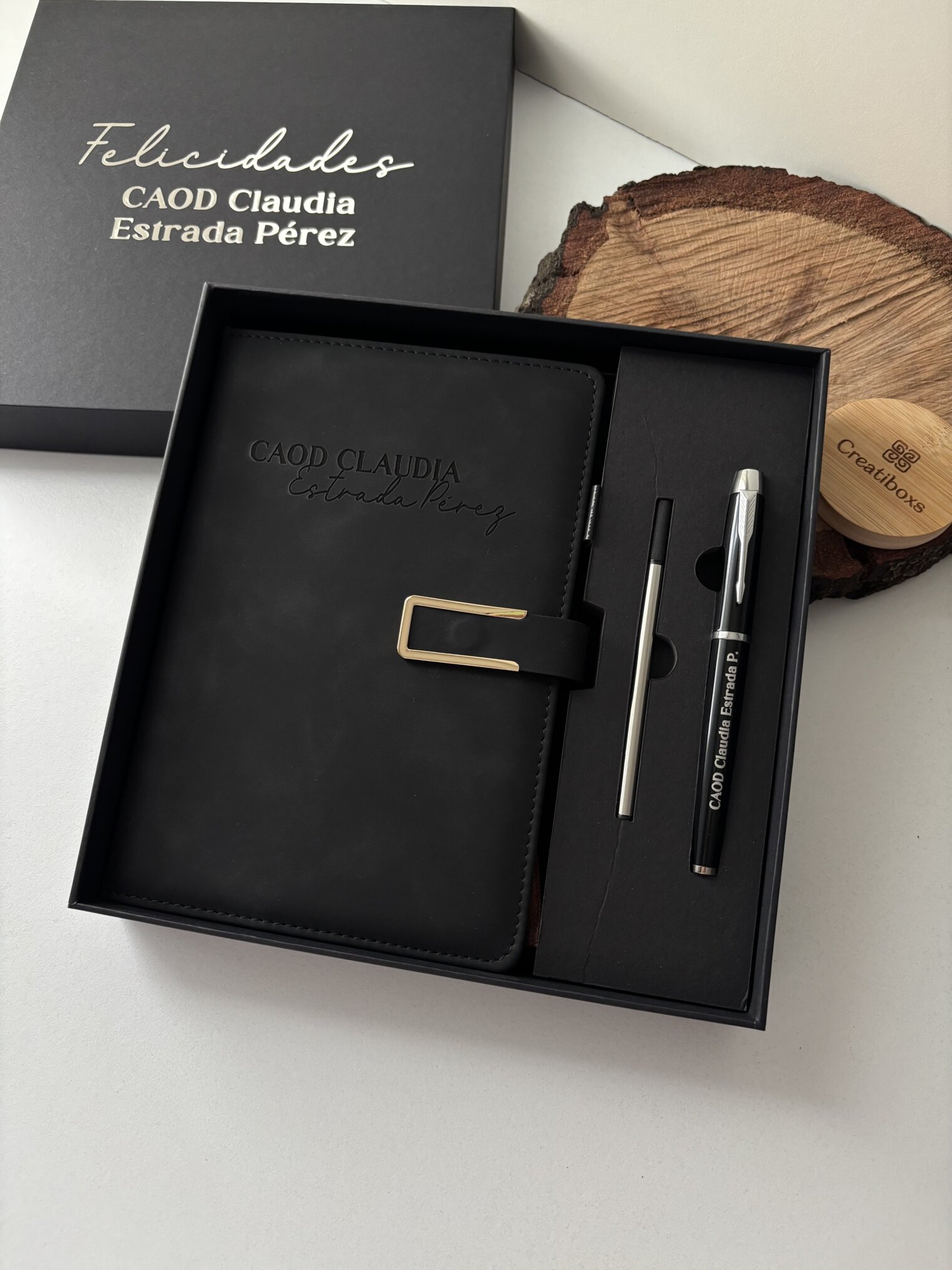 Caja de regalo negra con un Set libreta con broche magnético contorno metálico y bolígrafo en caja, además de un lápiz y un posavasos de madera sobre una superficie blanca.