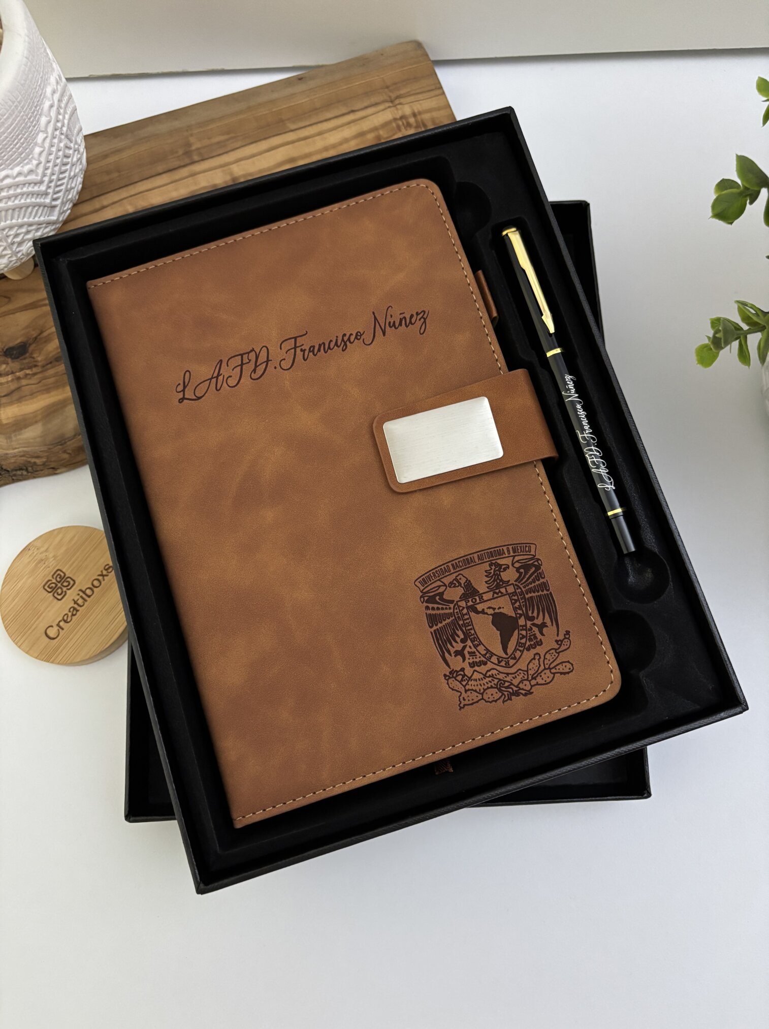 A Set libreta con broche magnético plateado y bolígrafo en caja negra, con grabado, colocado cerca de una planta y una bandeja de madera.