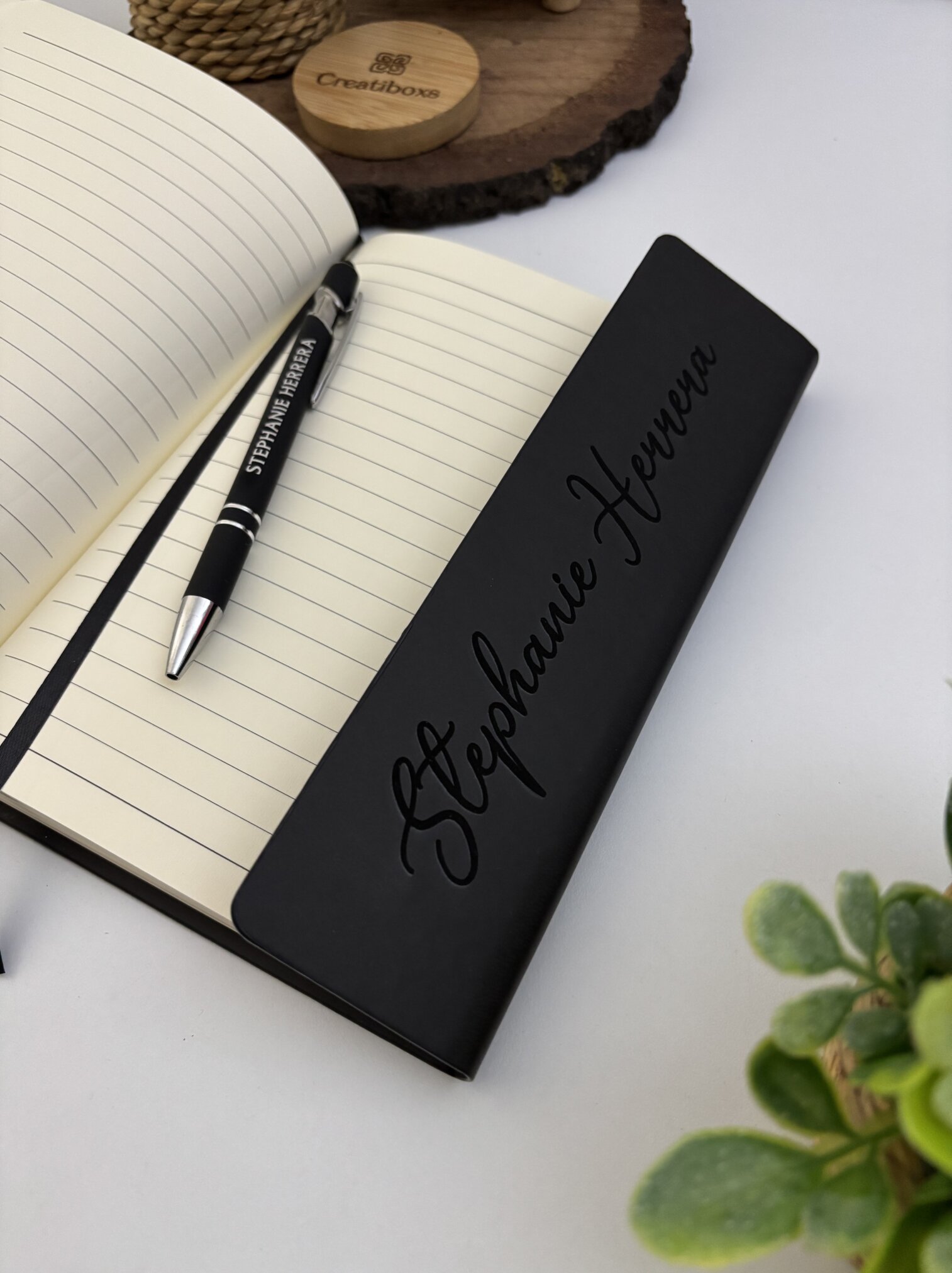 Un marcapáginas negro con Stephanie Herrera descansa sobre una "Libreta ejecutiva personalizada de pasta rígida alta calidad con cierre magnético vertical" abierta y un bolígrafo.