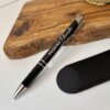 Un bolígrafo personalizado metálico con acabado brillante y detalles plateados, grabado con "PADRINO Eduardo", junto a un estuche negro sobre una tabla de madera.