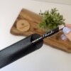Una mano sostiene un bolígrafo personalizado metálico con acabado brillante y detalles plateados sobre una tabla de madera con una pequeña planta.