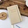 Una Mini libreta beige personalizada lisa con elástico con "Samantha Morales" en la portada descansa sobre una tabla de madera junto a una planta.