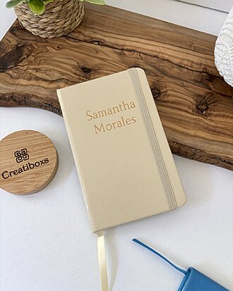 Una Mini libreta beige personalizada lisa con elástico con "Samantha Morales" en la portada descansa sobre una tabla de madera junto a una planta.