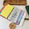 La Agenda planificador organigastos personalizada A6 broche magnético metálico se asienta sobre un escritorio blanco con coloridas pestañas, hojas de pegatinas y una bolsa transparente con la etiqueta Viajes, cerrada de forma segura con un cierre magnético.
