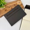 Una carpeta negra rotulada Arquitecto Abel Estrada con cierre magnético se encuentra junto a una Libreta ejecutiva personalizada de pasta rígida alta calidad con cierre magnético vertical, una planta y una tabla de madera sobre el escritorio.
