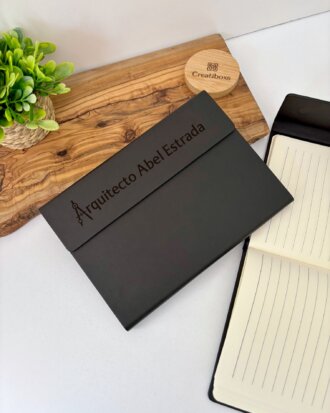 Una carpeta negra rotulada Arquitecto Abel Estrada con cierre magnético se encuentra junto a una Libreta ejecutiva personalizada de pasta rígida alta calidad con cierre magnético vertical, una planta y una tabla de madera sobre el escritorio.
