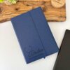 Libreta ejecutiva azul personalizada de pasta rígida alta calidad con cierre magnético vertical, con el nombre Doctor Guillermo Torres en la portada, junto a una planta y una bandeja de madera.