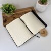 Dos "Libreta ejecutiva personalizada de pasta rígida alta calidad con cierre magnético vertical" abiertas descansan sobre un tablero de madera, con dos plantas en maceta y un posavasos redondo de madera cerca.