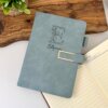 Una Libreta azul personalizada A5 con broche magnético contorno metálico, etiquetada Estefanía, descansa sobre una tabla de madera junto a un diario A5 forrado abierto y una planta en maceta.