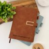 Libreta personalizada A5 con broche magnético contorno metálico, grabada con "Sergio Domínguez" en piel marrón, apilada sobre un cuaderno azul junto a plantas en un escritorio blanco.