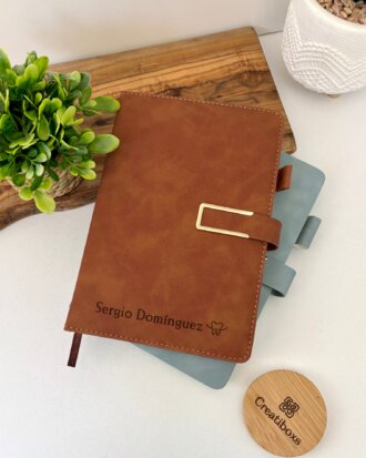 Libreta personalizada A5 con broche magnético contorno metálico, grabada con "Sergio Domínguez" en piel marrón, apilada sobre un cuaderno azul junto a plantas en un escritorio blanco.