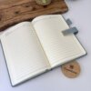 Una Libreta personalizada A5 abierta con broche magnético contorno metálico con páginas rayadas se encuentra sobre un escritorio, junto a una planta, una bandeja de madera y un posavasos Creatiboxes.