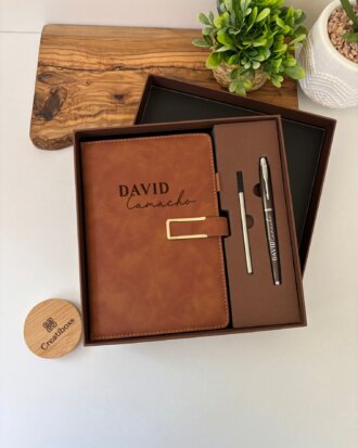 Set libreta con broche magnético contorno metálico y bolígrafo en elegante caja de regalo; fondo decorado con plantas.