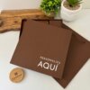 Set libreta con broche magnético contorno metálico y bolígrafo en caja marrón personalizada con "PERSONALIZA AQUÍ", junto a plantas y un objeto redondo de madera sobre superficie blanca.