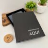 Caja de regalo negra con texto "PERSONALIZA AQUÍ", junto a un Set libreta con broche magnético plateado y bolígrafo en caja, colocado junto a plantas y una tapa de madera sobre una superficie clara.