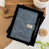 Set libreta con broche magnético plateado y bolígrafo en caja, personalizado con el nombre Lic. Javier Huerta acompañado de elementos decorativos como planta y acentos de madera. Javier Huerta, acompañado de elementos decorativos como planta y acentos de madera.