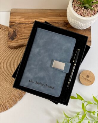 Set libreta con broche magnético plateado y bolígrafo en caja, personalizado con el nombre Lic. Javier Huerta acompañado de elementos decorativos como planta y acentos de madera. Javier Huerta, acompañado de elementos decorativos como planta y acentos de madera.