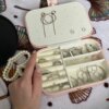 Una mano sostiene abierto un Joyero personalizado de cuero sintético chico con anillos, pendientes y una pulsera de perlas expuestos sobre una manta con motivos de conchas marinas.