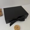 Una caja personalizada con 2 diseños magnética negra con cinta, junto a una tapa redonda de madera etiquetada Creatiboxs, sobre una superficie blanca.