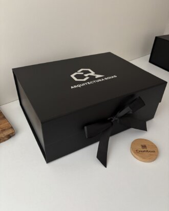 Una Caja negra personalizada con 2 diseños rígida magnética con un lazo y el logo ARQUITECTURA se asienta sobre una superficie blanca junto a una etiqueta redonda de madera, haciéndola perfecta para regalos especiales personalizados.