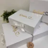 Caja personalizada blanca, rígida y magnética, con dos diseños, letras doradas, cintas blancas, una plantita y un círculo de madera.