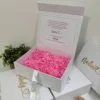 Caja personalizada con dos diseños, rígida y magnética, con papel rasgado de color rosa, junto a otras cajas y una pequeña planta verde.