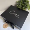 Una caja negra personalizada con dos diseños, rígida y con cierre magnético, adornada con una cinta negra, junto a una pequeña planta y una pieza de madera.