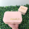 Una mano sostiene un Joyero personalizado de cuero sintético rosa chico etiquetado Marcela, con otra caja etiquetada Isabel que descansa sobre hojas verdes.