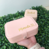 Una mano sostiene un joyero personalizado de cuero sintético chico en rosa con la etiqueta Marcela; otro igual, etiquetado Isabel, reposa sobre el follaje al fondo.