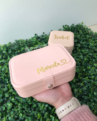 Una mano sostiene un joyero personalizado de cuero sintético chico en rosa con la etiqueta Marcela; otro igual, etiquetado Isabel, reposa sobre el follaje al fondo.