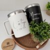 Dos tazas personalizadas etiquetadas como Cinthya Pastora y Mauricio Pastor se colocan en una bandeja de mimbre con una planta, ideal como regalo o combinadas con el Kit termo taza 360ml x2 y caja chica personalizada para momentos especiales.