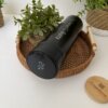El Termo personalizado digital liso 500ml (16oz) en negro muestra 27°C en su tapa digital, descansando sobre una bandeja tejida al lado de plantas en maceta. Este elegante termo mantiene sus bebidas a la temperatura ideal.
