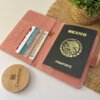 Un pasaporte mexicano en una funda rosa porta pasaporte personalizado con espacios para dinero y tarjetas seguridad RFID, junto a una planta y un objeto Creatiboxs redondo de madera, mantiene protegidos tus objetos esenciales.