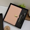 Un Set agenda rosa personalizado con broche rectángulo dorado y bolígrafo en caja en un elegante estuche de regalo negro, expuesto junto a un posavasos de madera Creatiboxs y una planta verde.