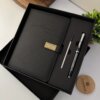 Set agenda personalizada con broche rectángulo dorado y bolígrafo en caja, color negro 2026, acompañado de un lápiz. Presentado junto a una planta y un portavasos de madera.
