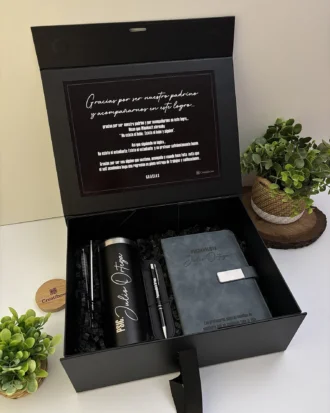 Kit regalo personalizado en caja negra con termo delgado 590ml, libreta broche metálico y bolígrafo, ideal para cualquier ocasión. Incluye nota de agradecimiento y se presenta rodeado de plantas verdes para un toque elegante.