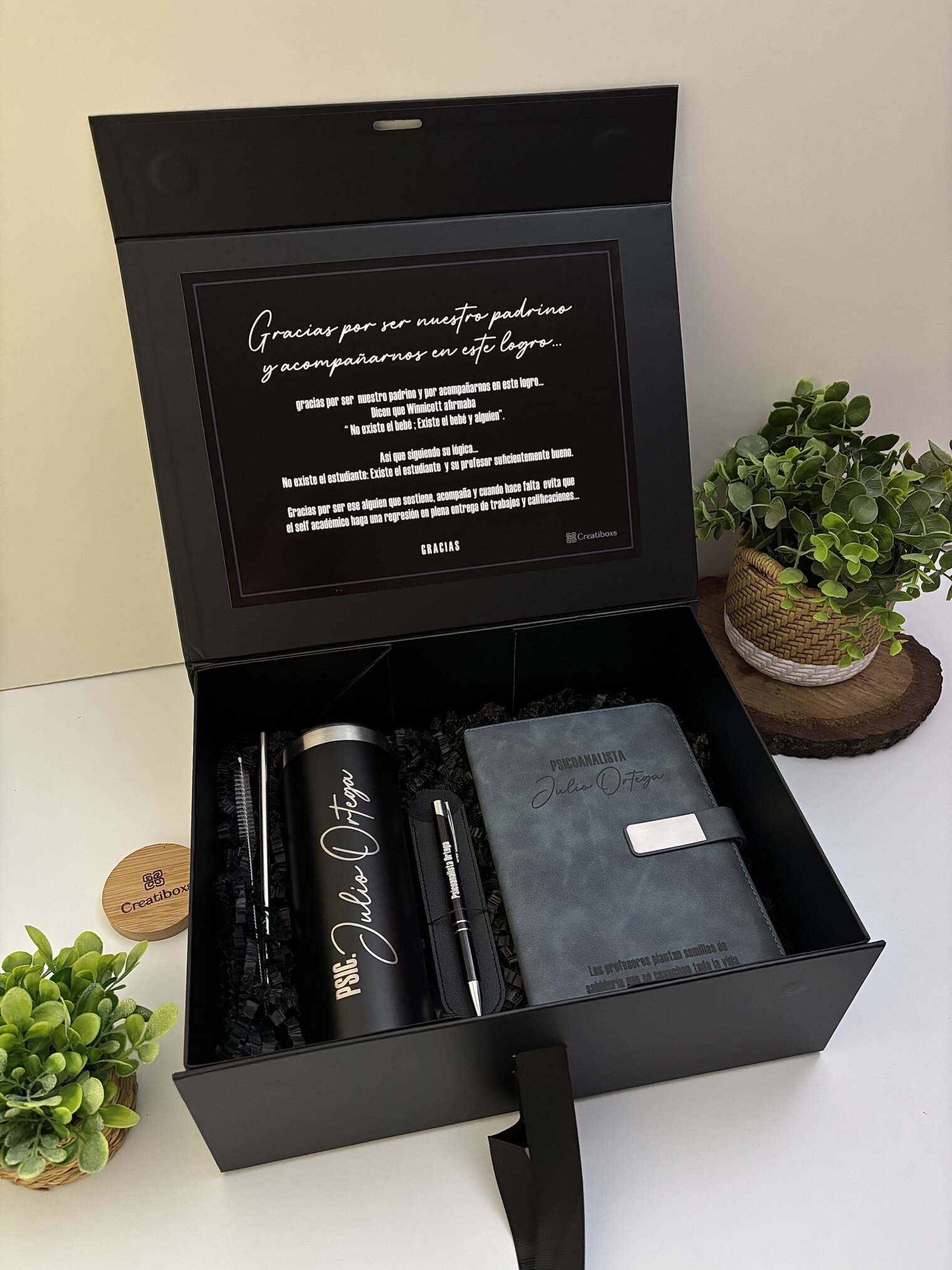 Kit regalo personalizado en caja negra con termo delgado 590ml, libreta broche metálico y bolígrafo, ideal para cualquier ocasión. Incluye nota de agradecimiento y se presenta rodeado de plantas verdes para un toque elegante.
