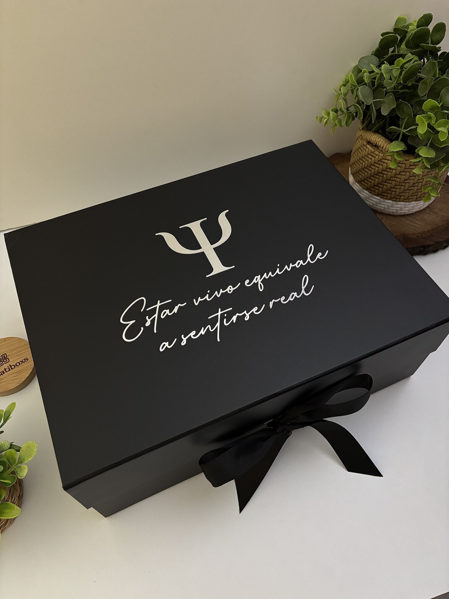 Kit regalo personalizado en caja negra con cinta, incluye termo delgado 590ml, libreta con broche metálico y bolígrafo. Ideal para cualquier ocasión y presentado junto a plantas verdes sobre superficie blanca.