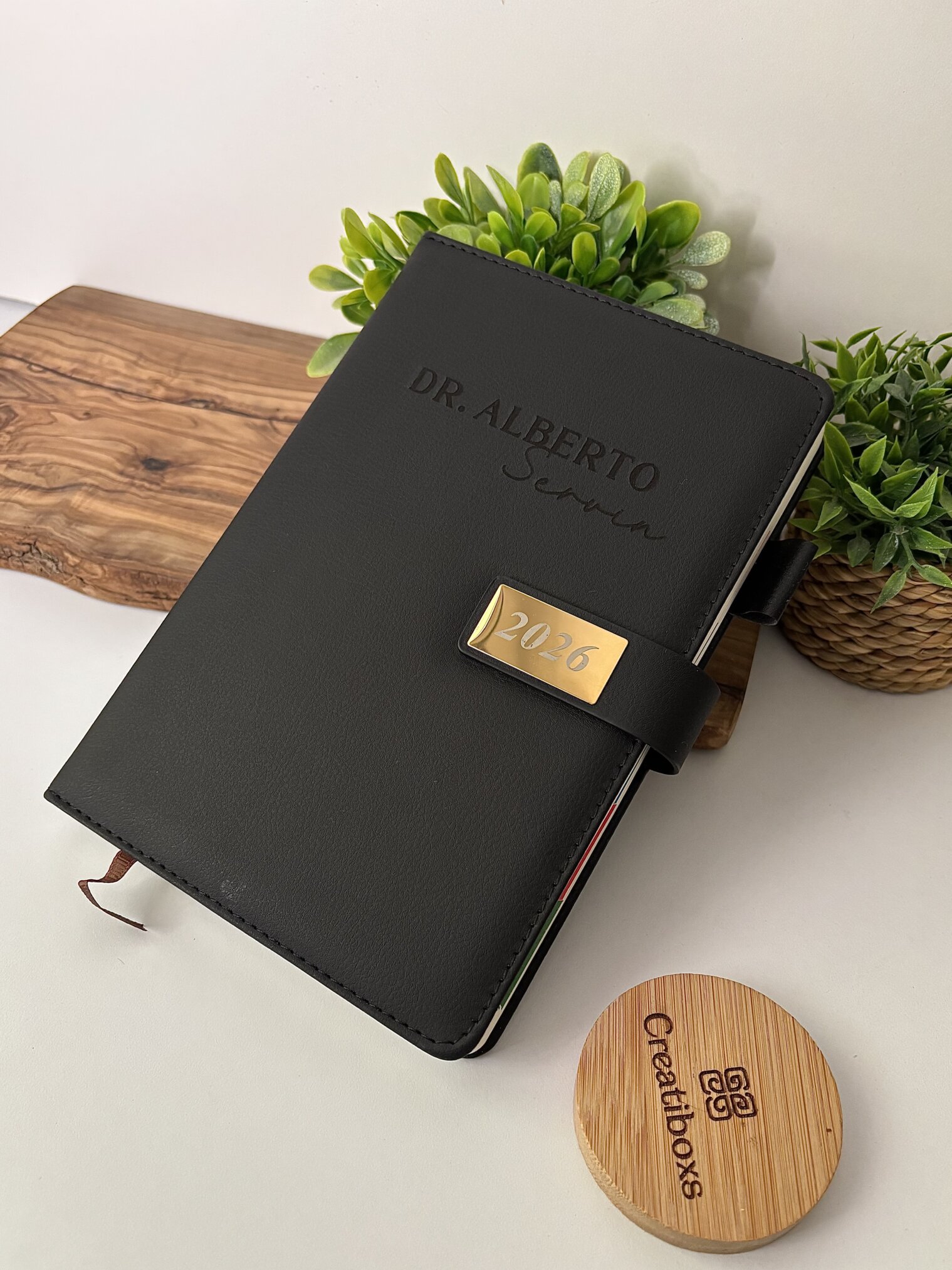 A black "Agenda personalizada A5 con broche magnético rectángulo dorado" rotulada Dr. Alberto descansa sobre una tabla de madera, con plantas verdes al fondo.