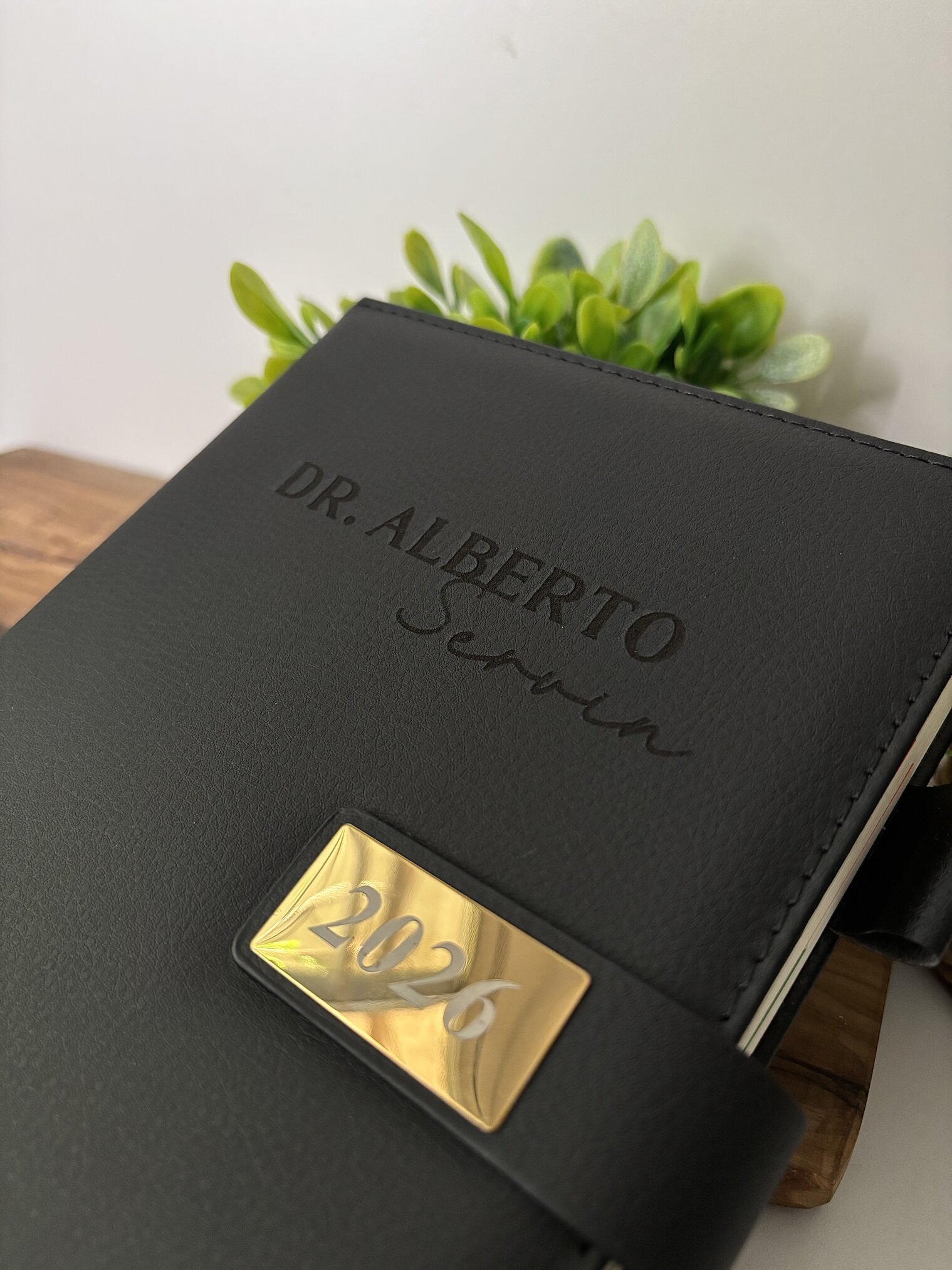 Agenda personalizada A5 con broche magnético rectángulo dorado 2026, negra, personalizada para Dr. Alberto Servin, junto a hojas verdes.