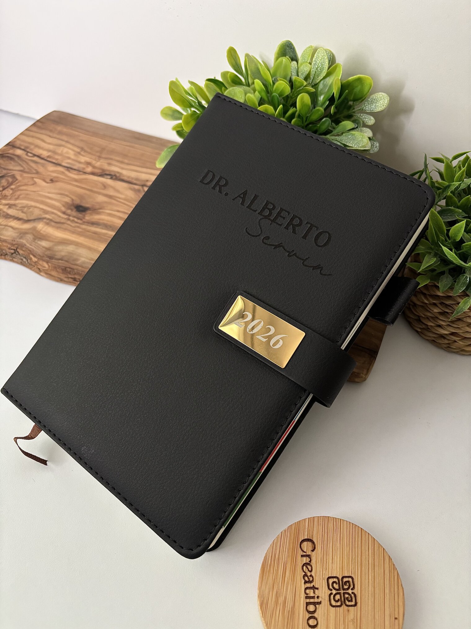 Una "Agenda personalizada A5 con broche magnético rectángulo dorado" rotulada Dr. Alberto descansa sobre una tabla de madera junto a una planta verde.