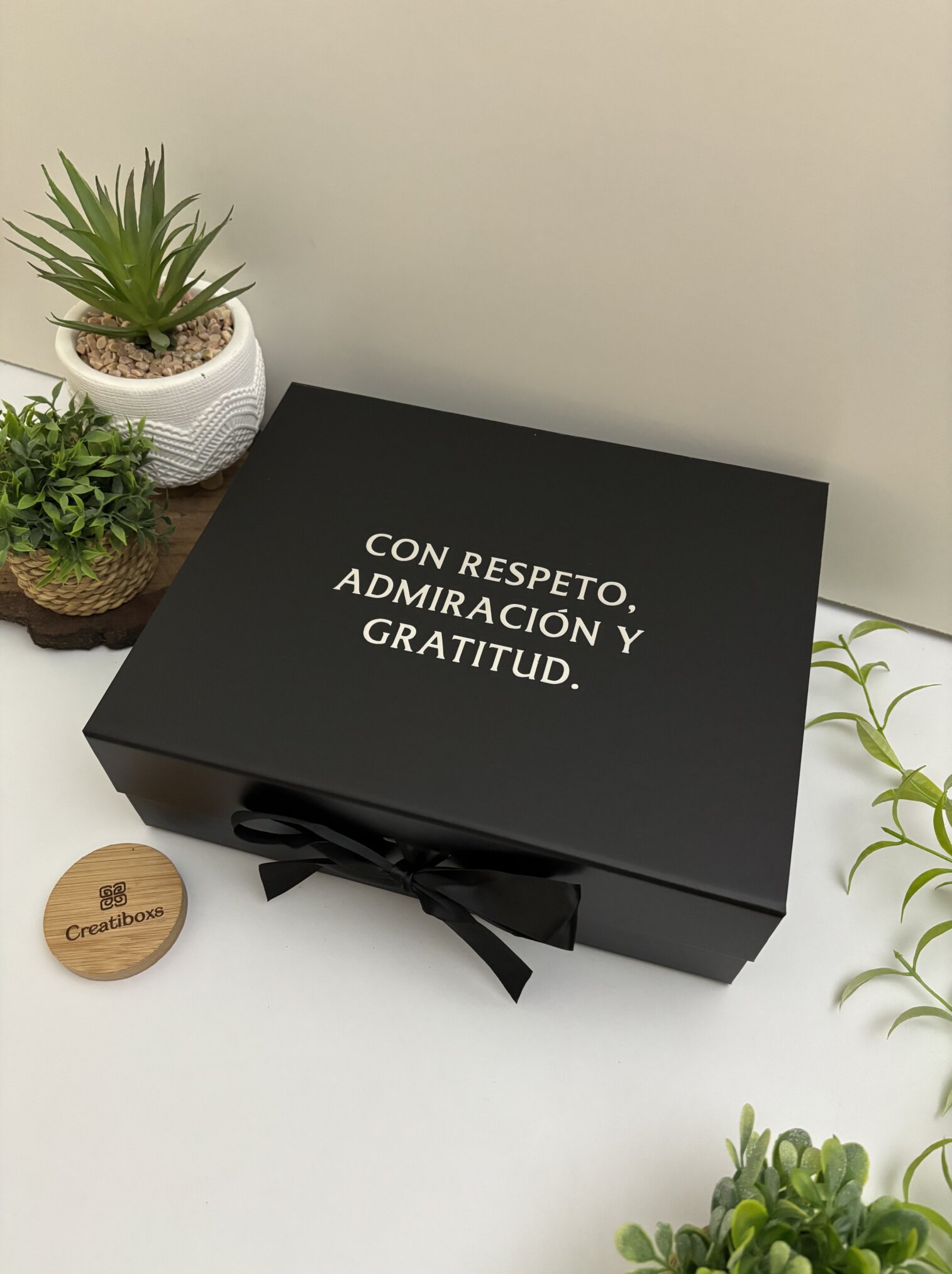 Kit regalo personalizado en caja con termo delgado 590ml, libreta con broche metálico y bolígrafo, presentado sobre superficie blanca con plantas verdes alrededor.