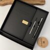 Set negro agenda personalizada con broche rectángulo dorado y bolígrafo en caja, bellamente presentado junto a una planta y tapa de madera.