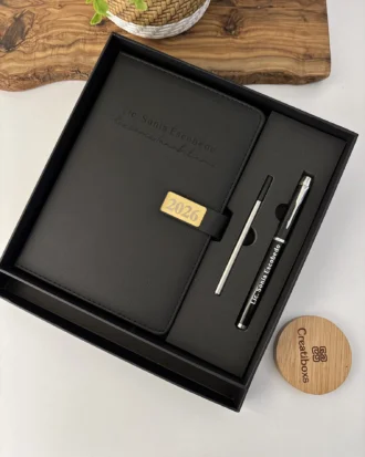 Set negro agenda personalizada con broche rectángulo dorado y bolígrafo en caja, bellamente presentado junto a una planta y tapa de madera.