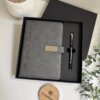Set libreta con broche magnético plateado y bolígrafo negro en caja, acompañado de una pequeña planta y un portavasos Creatibks sobre la mesa.