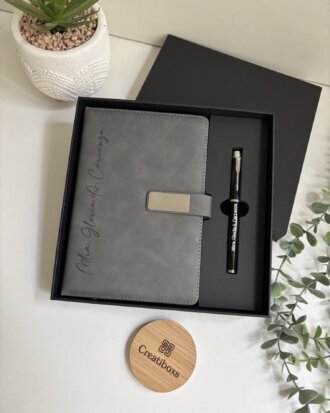 Set libreta con broche magnético plateado y bolígrafo negro en caja, acompañado de una pequeña planta y un portavasos Creatibks sobre la mesa.